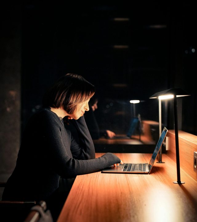 Focused woman using laptop at night in a dimly lit workspace, emphasizing concentration and technology.